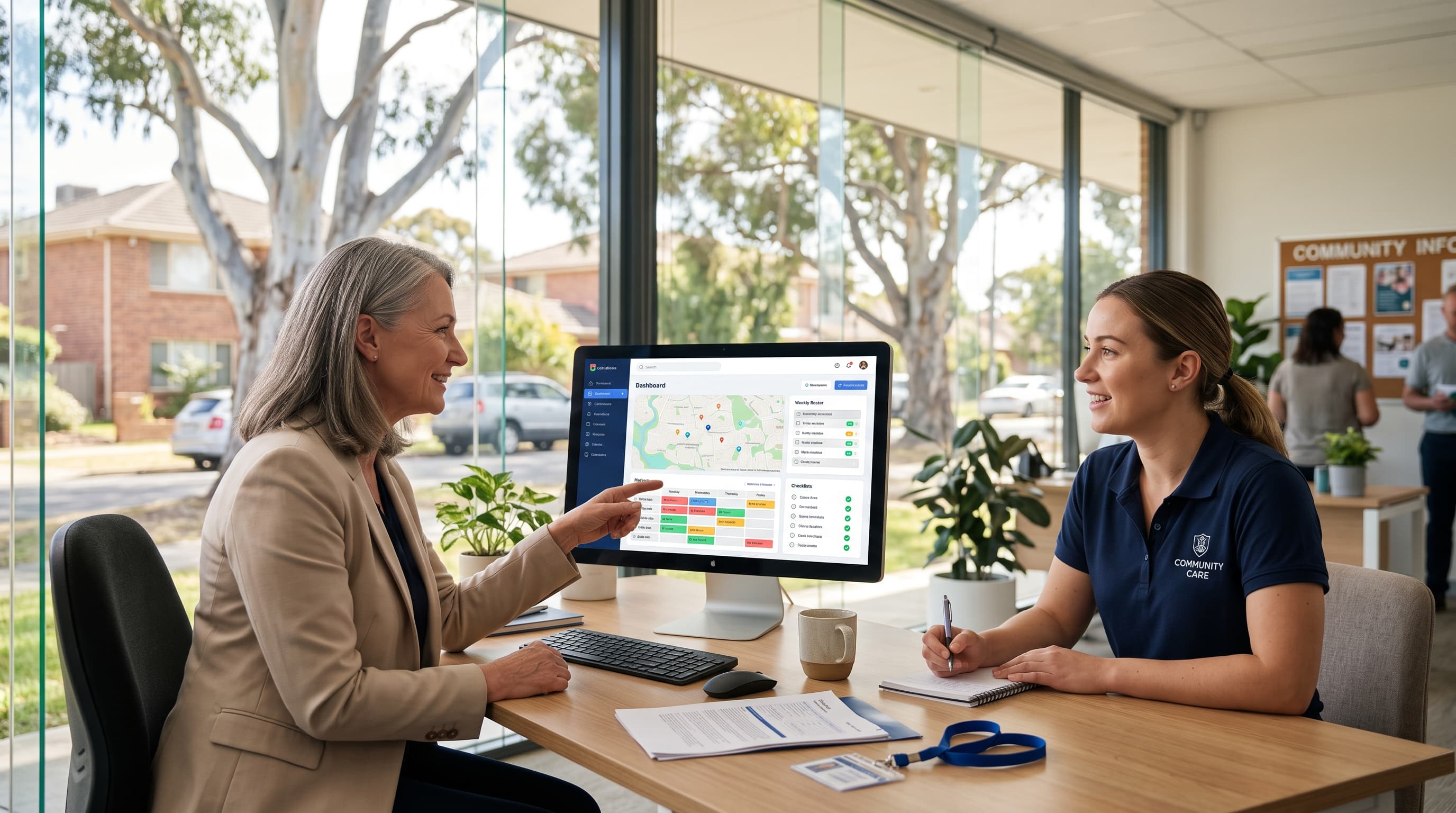 Two women talk across a desk, one pointing to a roster dashboard on the monitor